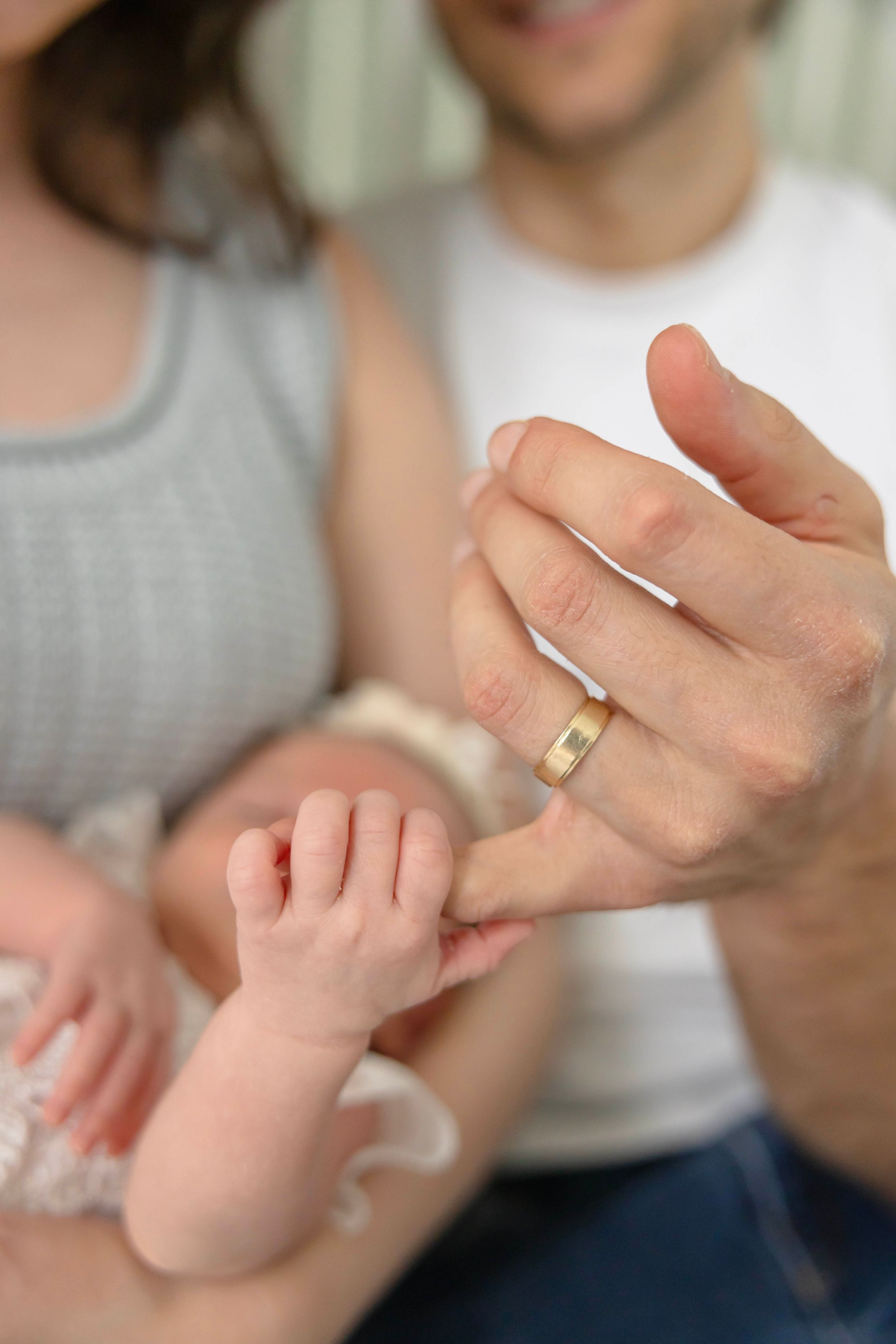 A newborn baby grasping the finger of a father wearing a gold wedding ring, held by parents in a tender family moment.