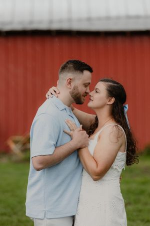 Couple embracing near a red barn, woman in white dress with blue bow, man in light blue shirt, sharing a romantic moment.