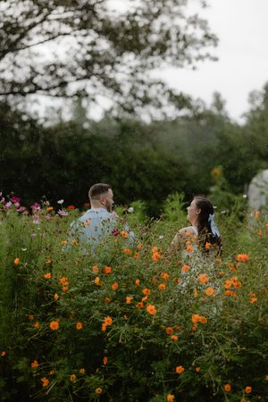 Couple standing together in a lush wildflower garden with orange cosmos blooms in the foreground.