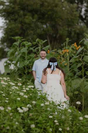 Couple walking through sunflower garden, woman in white dress with blue bow, man in light blue shirt smiling.