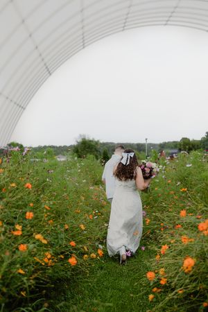 Bride walks through colorful wildflower field in white gown holding bouquet under large white canopy structure.