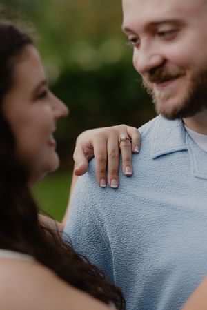 Engaged couple smiling at each other, woman resting hand with diamond engagement ring on man's shoulder.