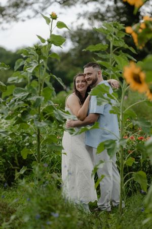 Couple embracing among tall sunflowers in a lush garden, woman in white dress smiling at camera.