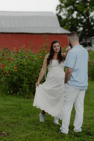 Couple dancing in white outfits near colorful flower garden with red barn backdrop on green grass.