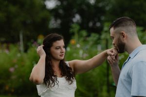 A man kisses a woman's hand outdoors in a lush green garden setting.
