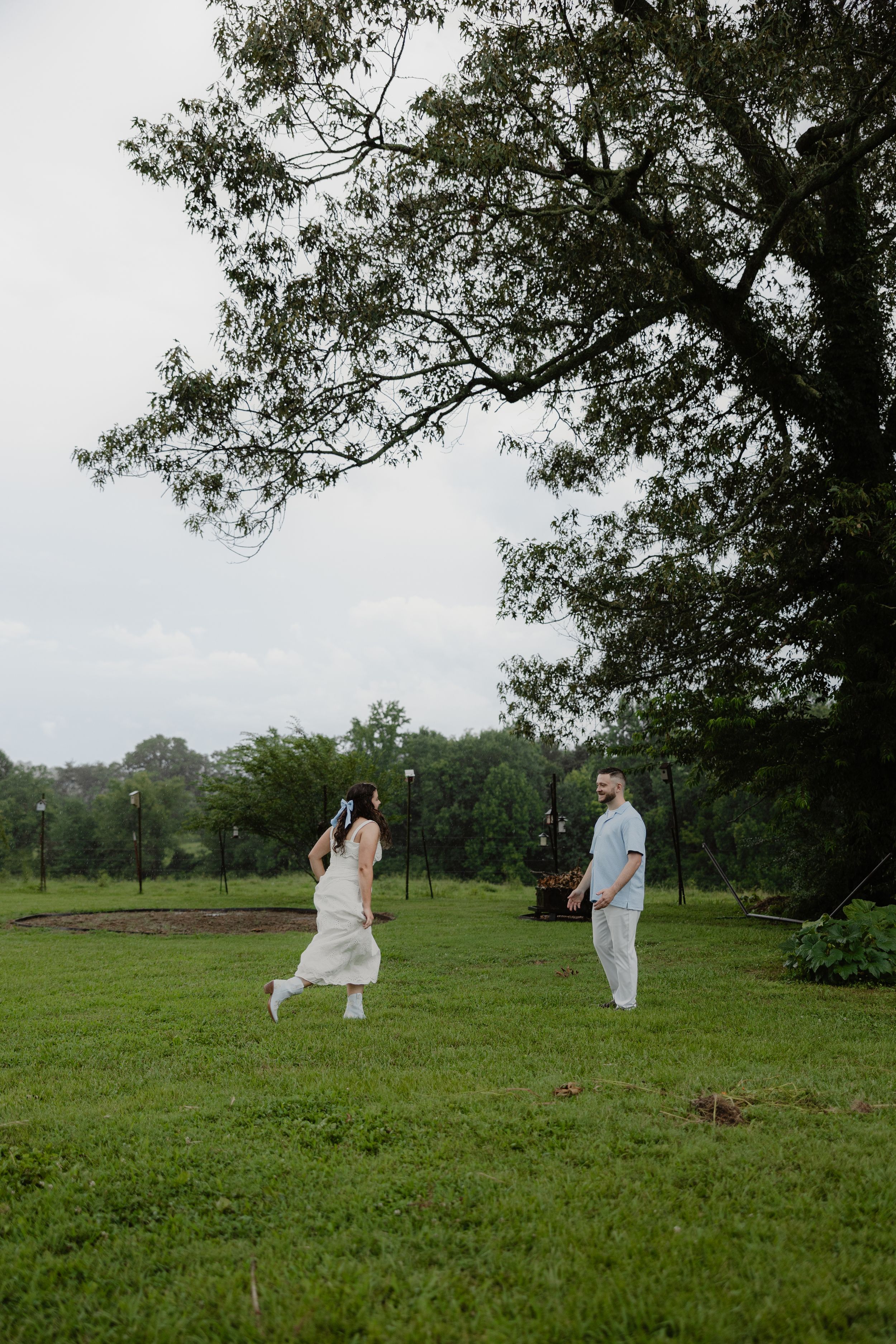 Couple running toward each other on lush green lawn beneath large tree on cloudy day.