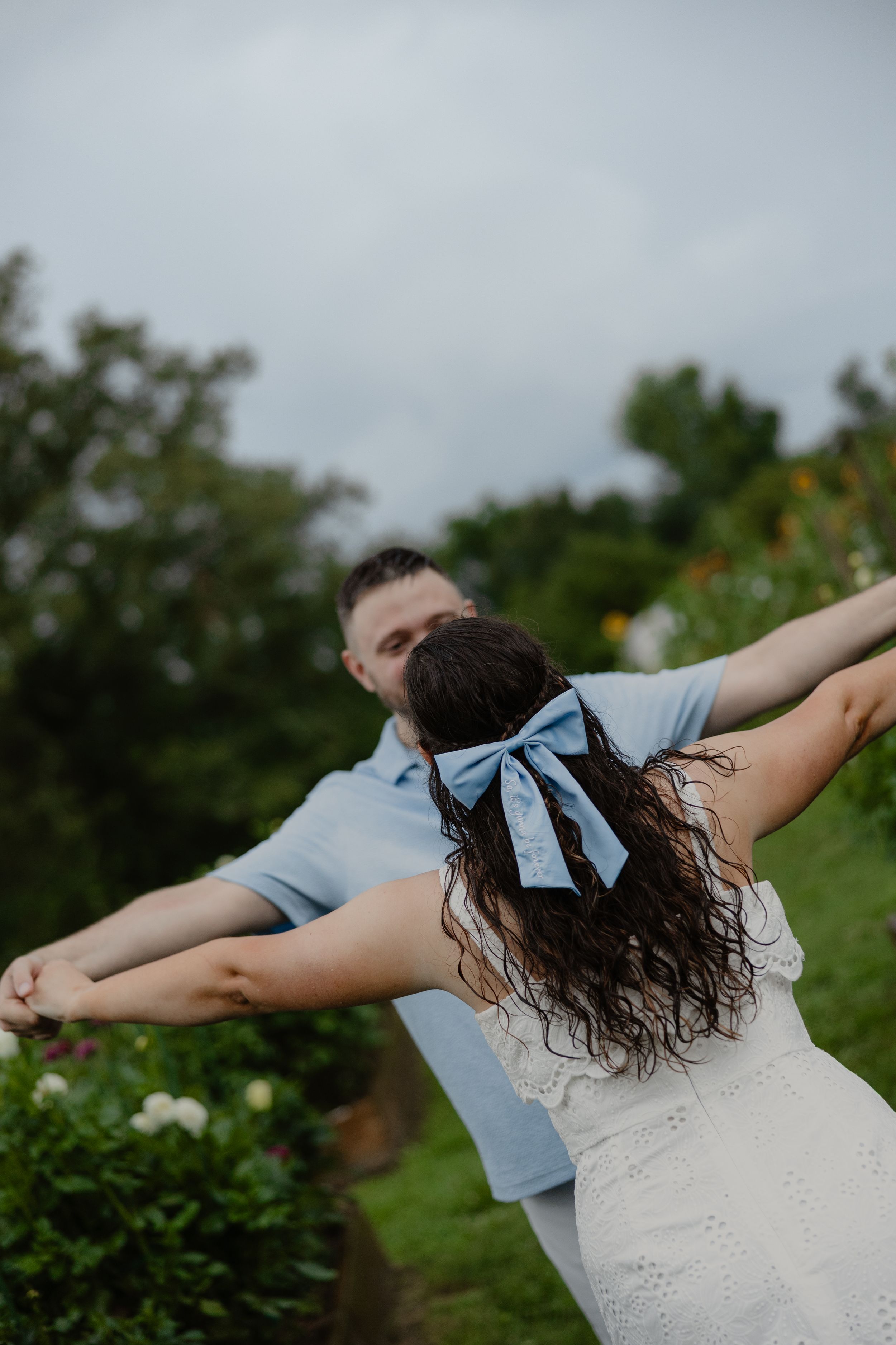 Couple dancing outdoors, woman with blue bow in dark curly hair wearing white dress, man in blue polo shirt, garden background.