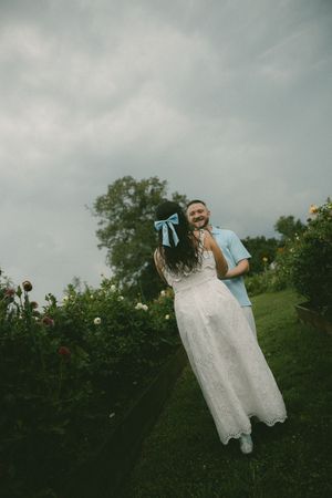 Joyful couple dancing in a flower garden, woman wearing white lace dress with blue bow in her hair.