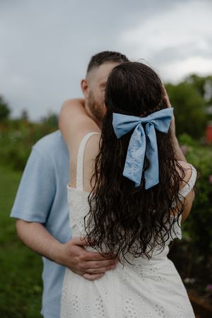 Couple embracing outdoors, woman with dark curly hair adorned with a blue bow, wearing a white dress.