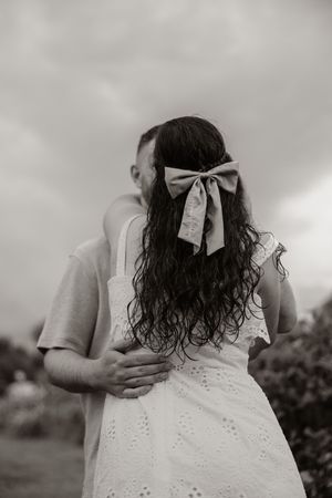 Black and white photo of couple embracing outdoors, woman in white eyelet dress with bow in curly hair.