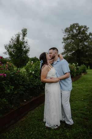 Couple sharing a romantic kiss in a lush garden, bride in white lace dress and groom in light blue outfit.