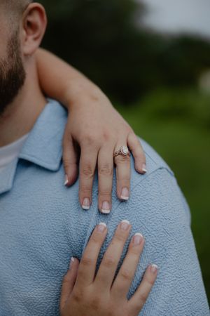 Close-up of a woman's hands with French manicure and engagement ring resting on a man's blue jacket shoulder.
