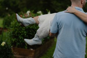 A man in a blue shirt carries a child in a white dress outdoors near garden beds.