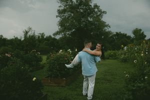 Man lifting woman in embrace among garden flowers under dramatic stormy sky.