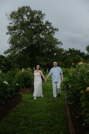 Smiling bride and groom hold hands walking through a lush flower garden on a cloudy day.