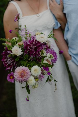 Bride in white dress holding lush purple and white dahlia wedding bouquet with greenery accents.