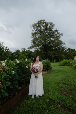 Bride in white lace dress holding pink bouquet stands in lush green garden with flowers and large tree backdrop.