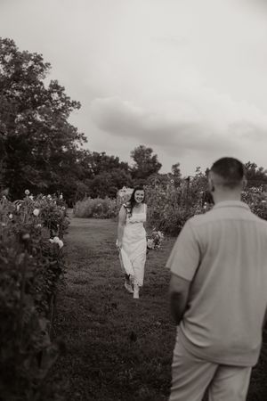 Black and white photo of bride in white dress walking toward groom through a lush garden path on cloudy day.