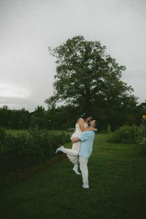 Man lifting woman in white dress outdoors, kissing near large green tree on overcast day.