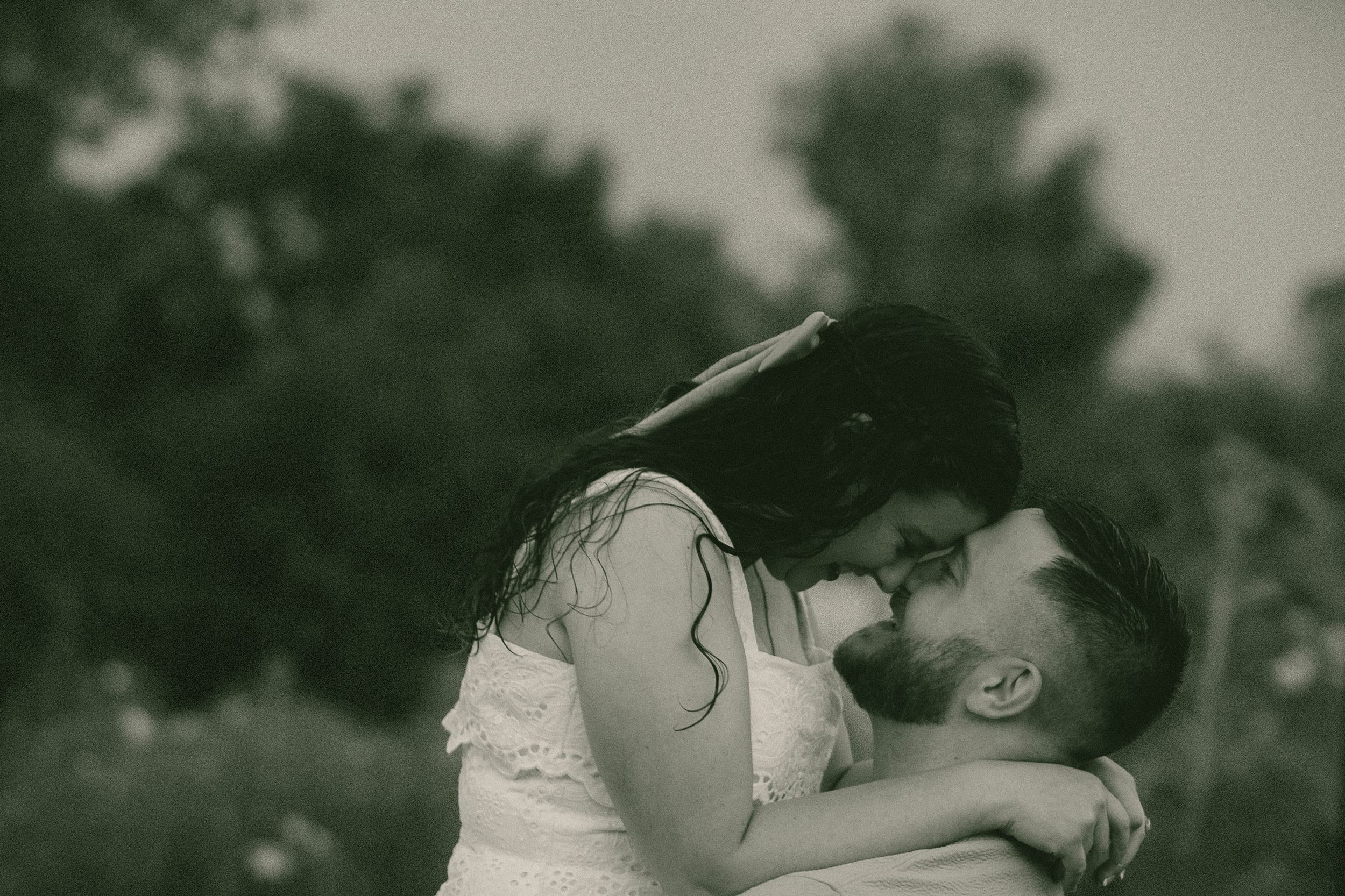 Romantic black and white photo of couple sharing a tender kiss outdoors surrounded by soft bokeh trees.