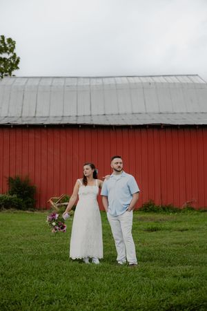 Bride in white lace dress and groom in blue shirt hold hands before a red barn on green grass.