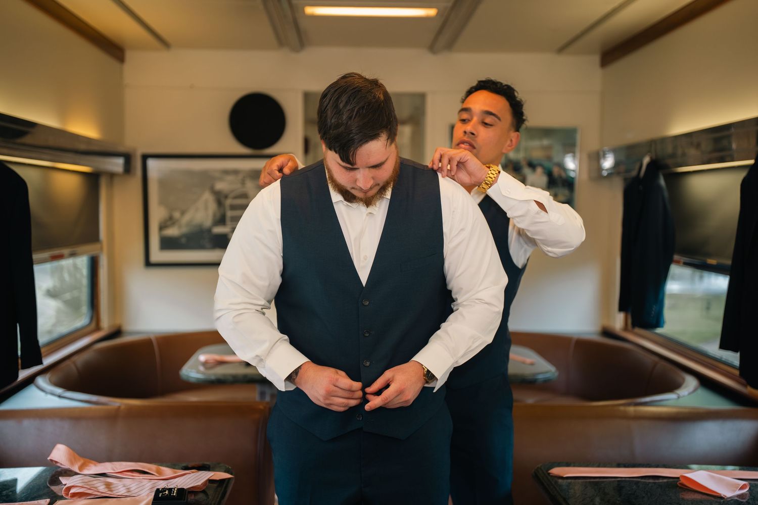 Groom buttoning vest while groomsman adjusts his collar in an elegant dressing room before wedding.