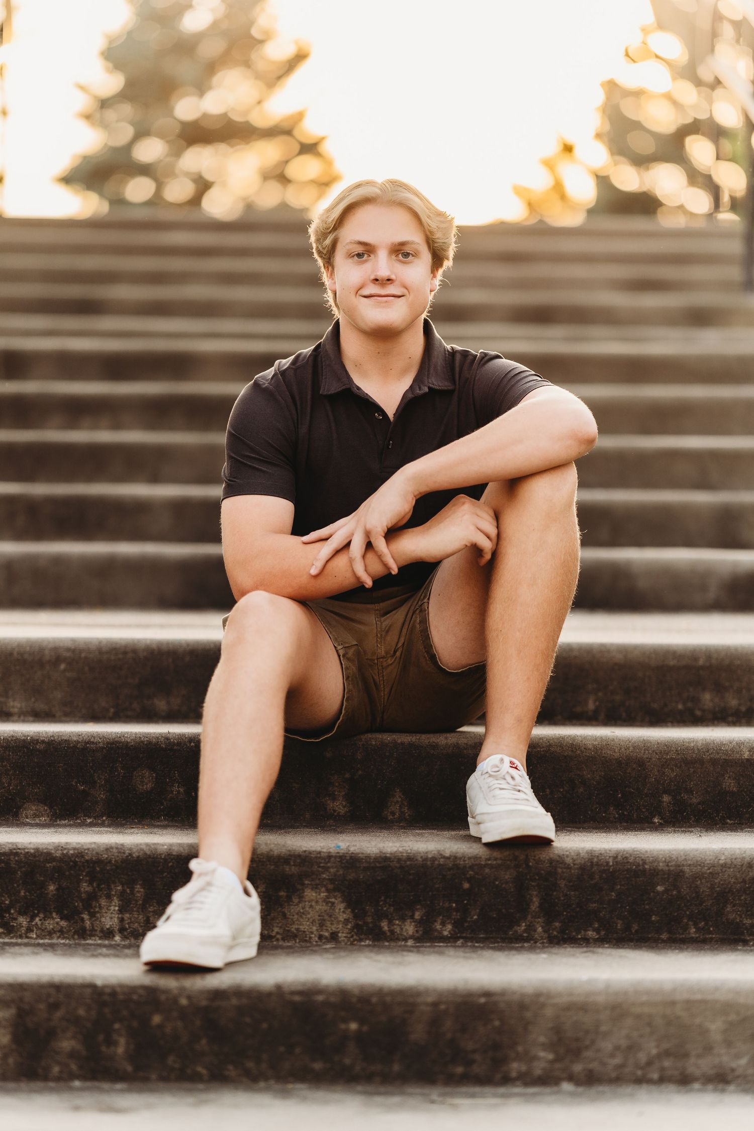 Young man in black polo and khaki shorts sitting on stone steps outdoors during golden hour sunset.
