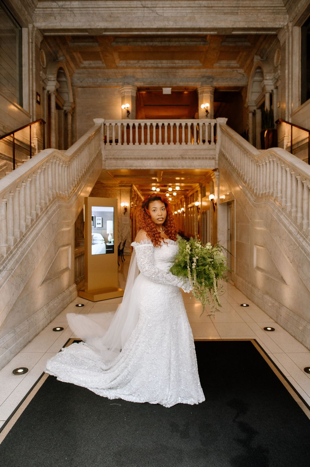 Bride in white lace off-shoulder gown holding greenery bouquet stands between grand marble staircases in elegant venue.
