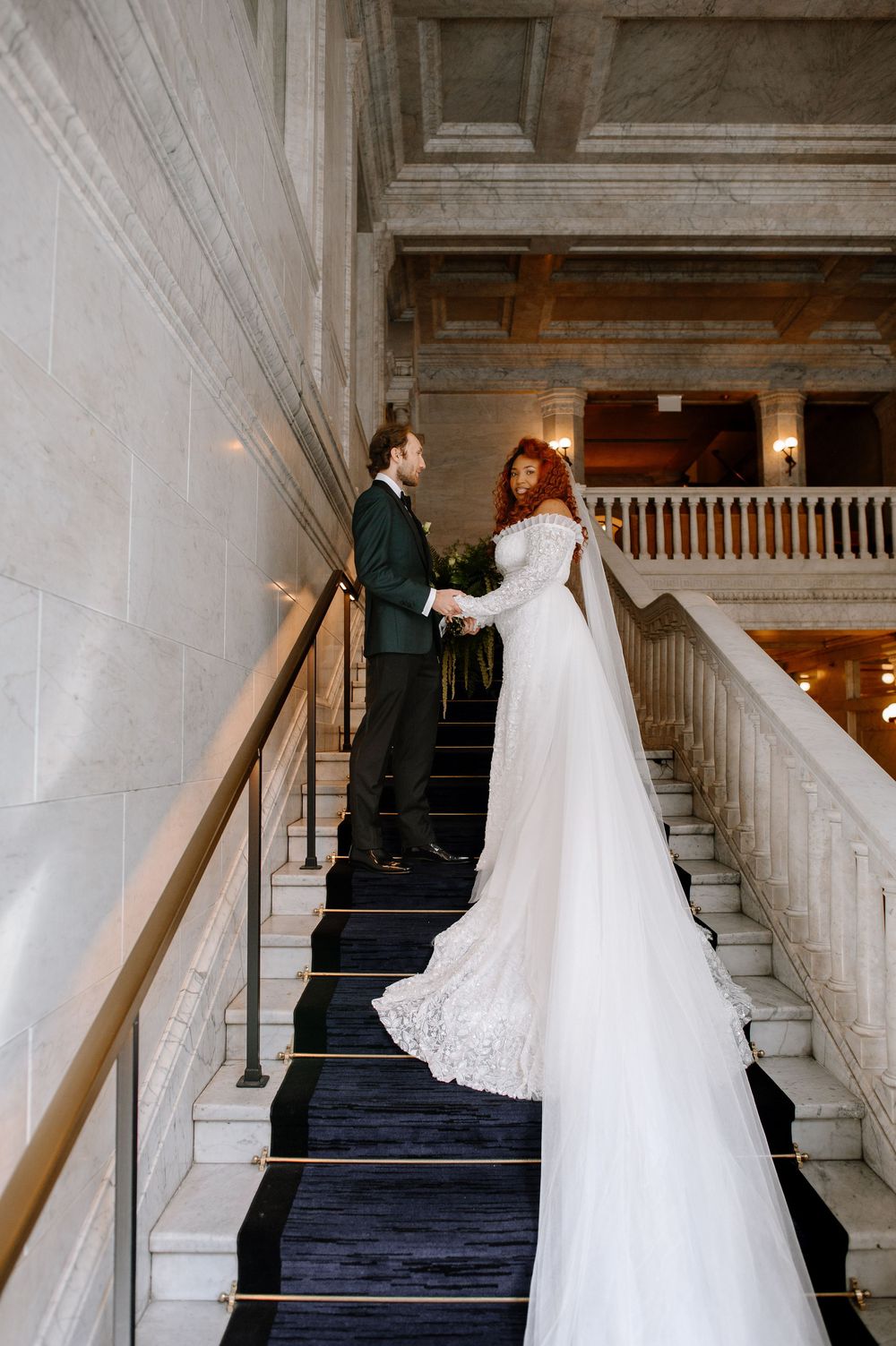 Bride in lace gown with veil and groom in dark suit hold hands on grand marble staircase in elegant venue.