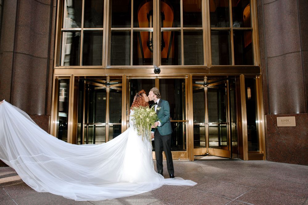 Bride and groom share a romantic kiss outside elegant gold revolving doors, her cathedral veil flowing dramatically.