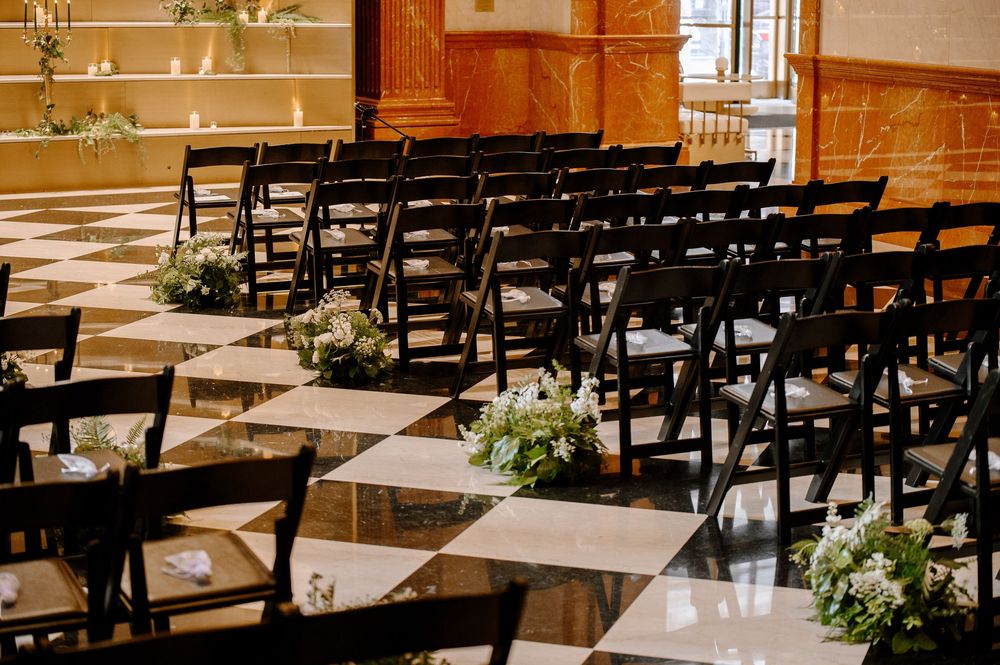 Elegant wedding ceremony setup with dark folding chairs on checkered marble floor adorned with white floral arrangements.