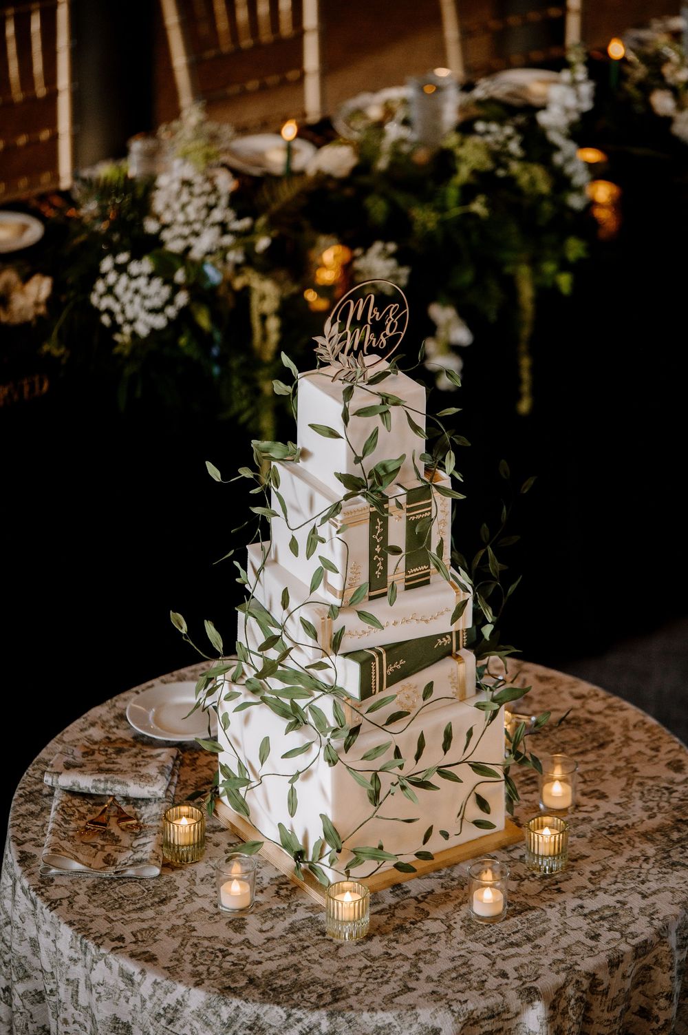 Elegant four-tier square wedding cake with green leaf details and Mr. & Mrs. topper, surrounded by candles on lace tablecloth.