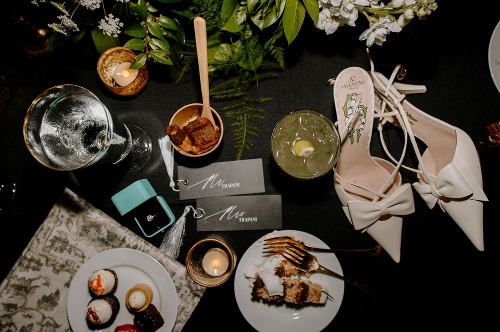 Elegant wedding flat lay with white heels, cocktails, desserts, a ring box, and place cards on a dark table.