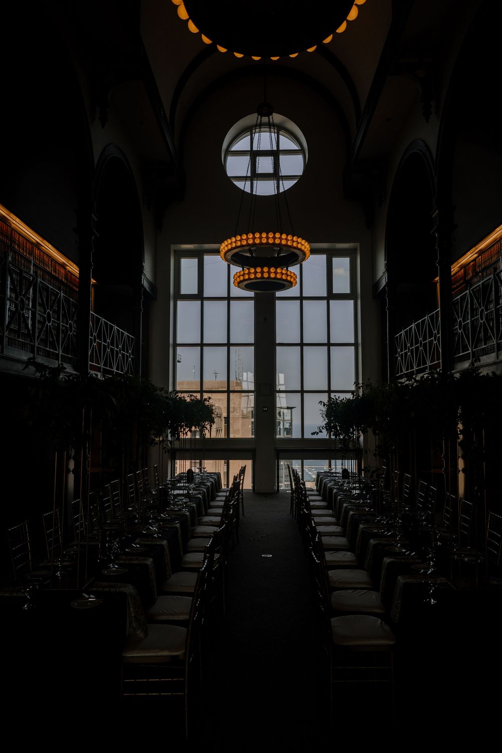 Elegant dark banquet hall with glowing chandelier, arched windows, and symmetrical dining tables set for a formal event.