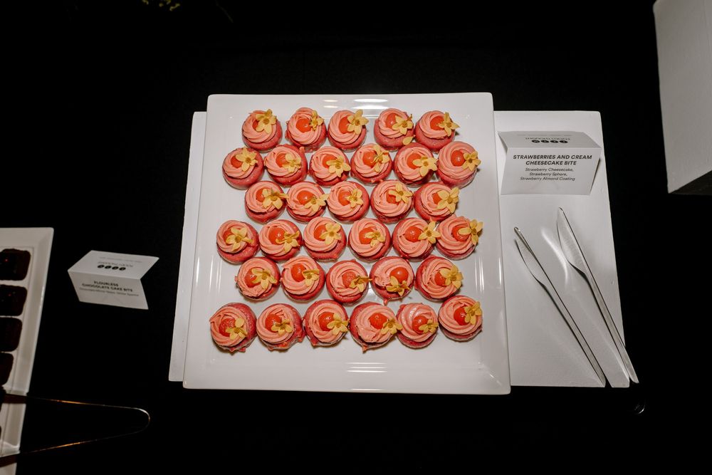 A white tray of pink frosted cupcakes with orange swirl decorations displayed on a dark table at a catered event.