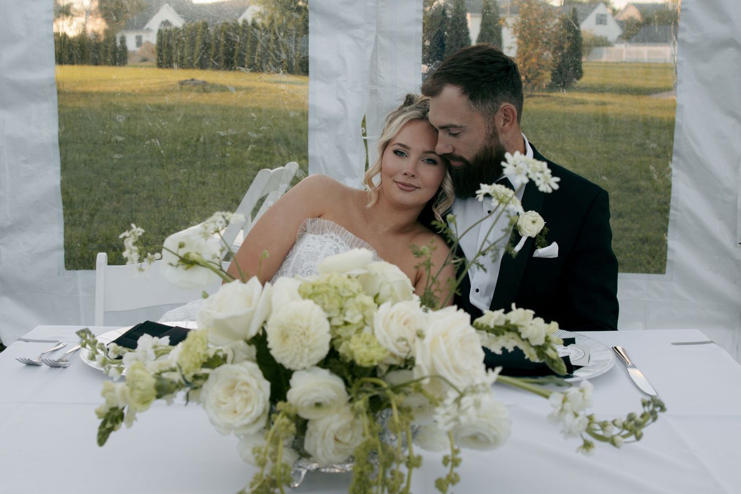 Elegant outdoor event with guests in black attire gathered near a white marquee tent on a sunny evening.