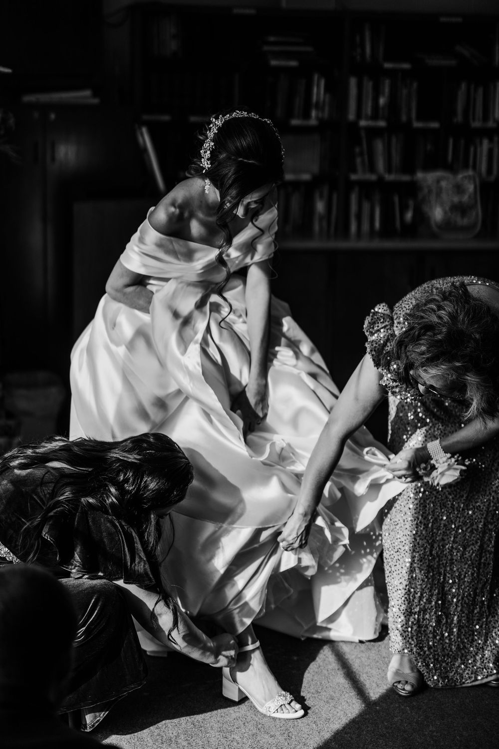Bride in elegant white gown gets shoes put on by helpers in a dramatic black and white photo.