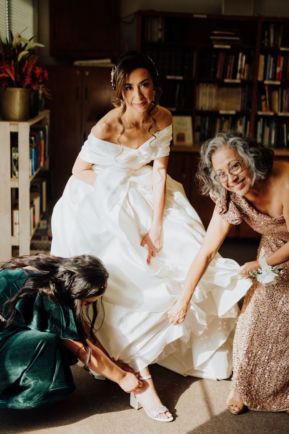 Bride in off-shoulder ball gown gets shoes fastened by two women in a sunlit library setting.