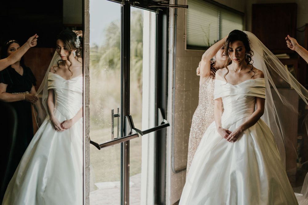 Bride in off-shoulder white ball gown with veil reflected in glass doors before her wedding.