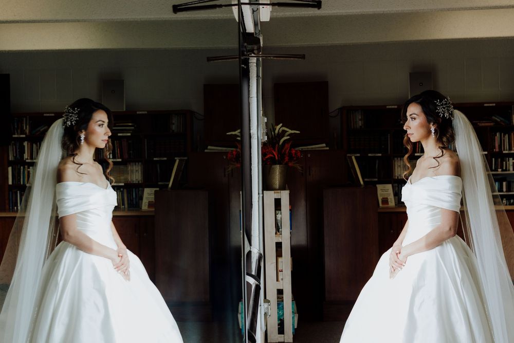 Bride in white off-shoulder ball gown reflected in mirror surrounded by bookshelves.