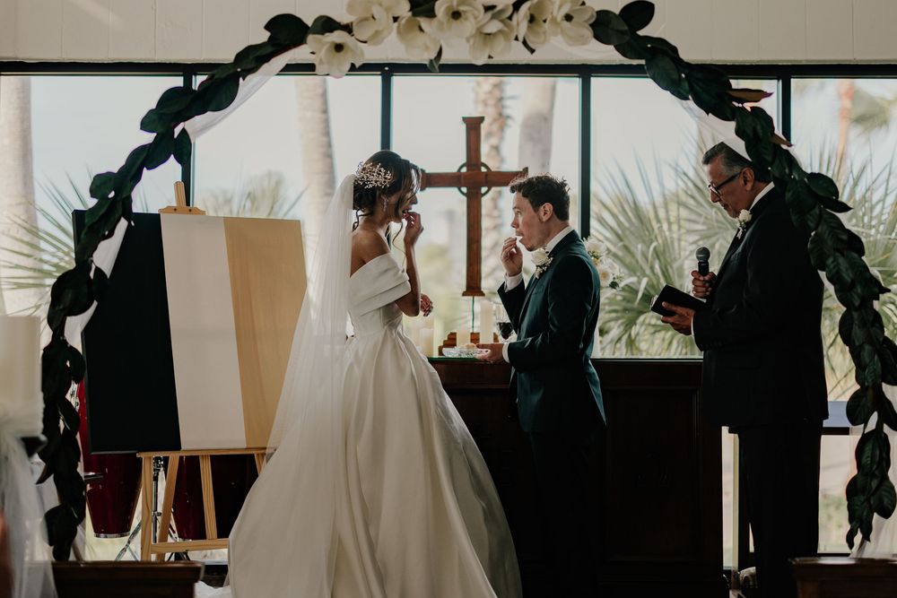 Bride and groom exchanging vows at church altar beneath floral arch with cross.