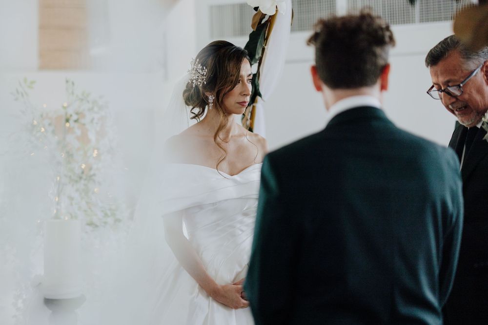 Bride in off-shoulder white gown gazing at groom during intimate wedding ceremony.
