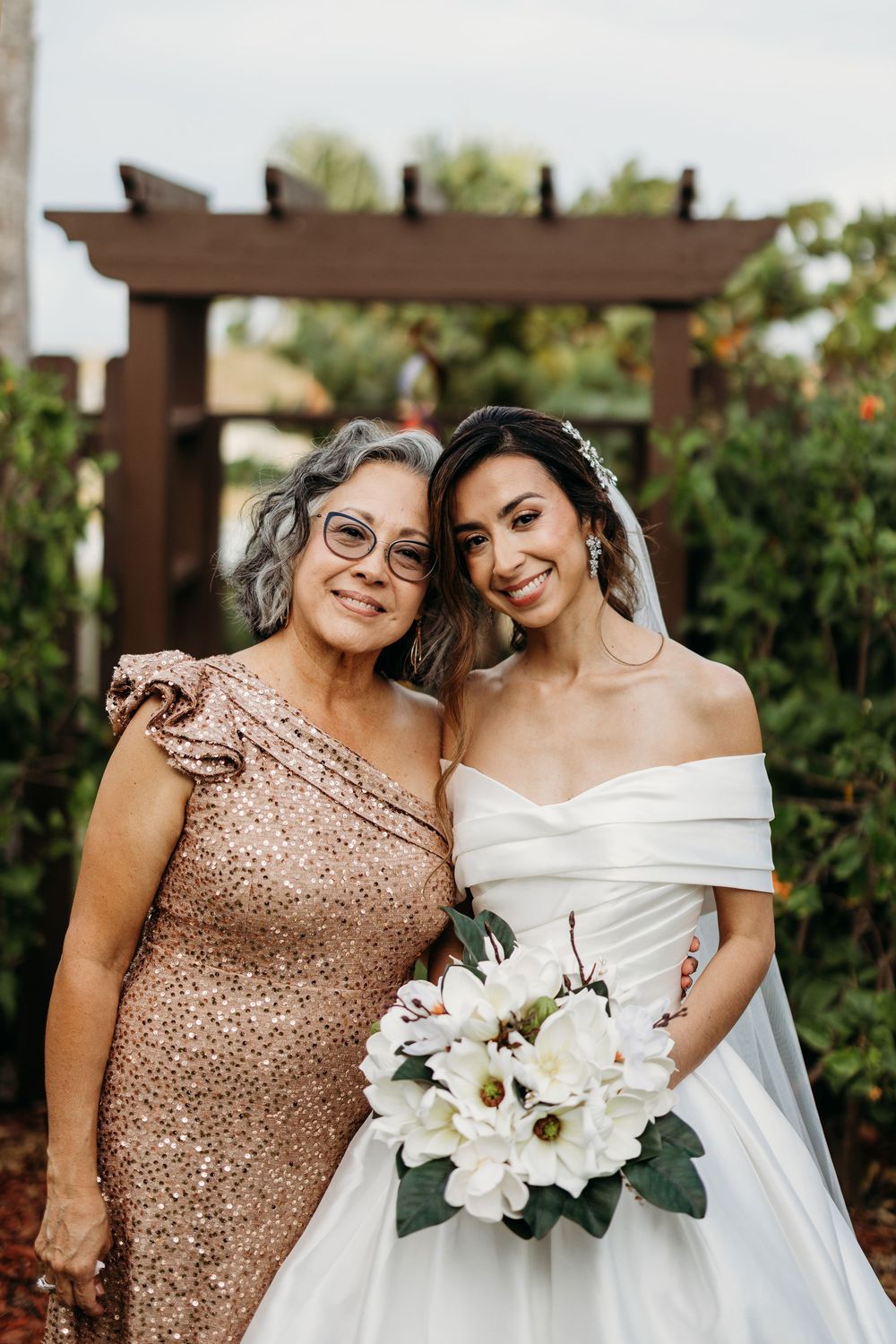 Bride in white off-shoulder gown poses with older woman in rose gold dress.