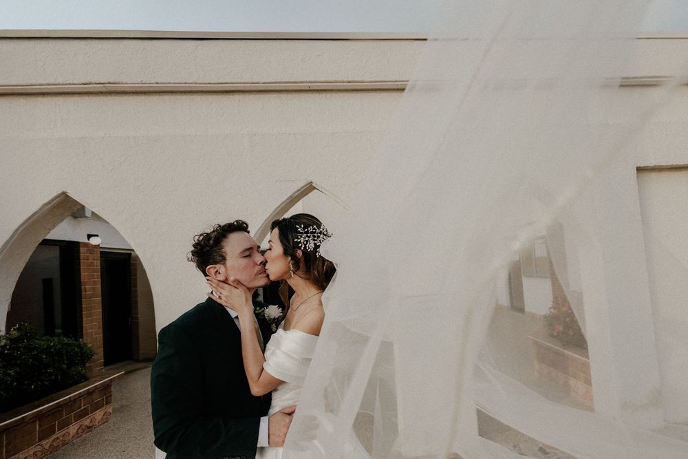 Bride and groom share a kiss outdoors as wedding veil billows in the wind.