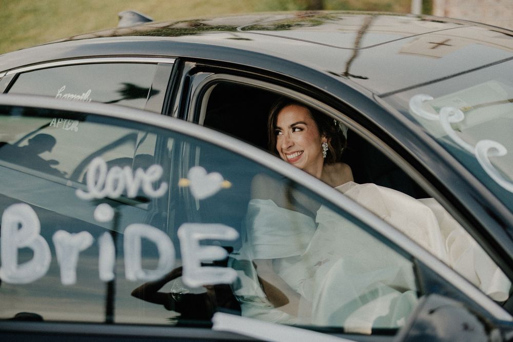 Smiling bride in white gown sits in car decorated with wedding messages.