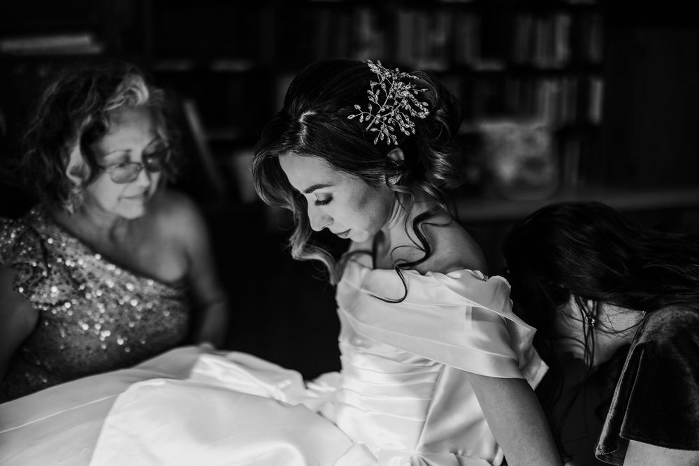 Bride in off-shoulder white gown gets assisted by family members in a black and white photo.