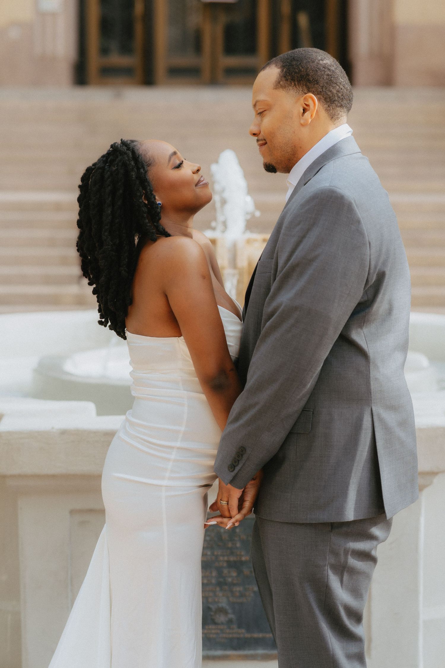 A couple in wedding attire holds hands by a fountain, the bride in white gown gazing up at the groom in a gray suit.