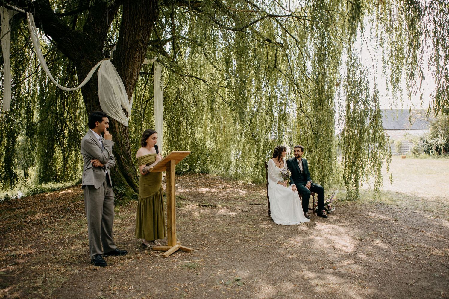 Outdoor wedding ceremony under a weeping willow tree with bride, groom, officiant, and guest in a natural garden setting.
