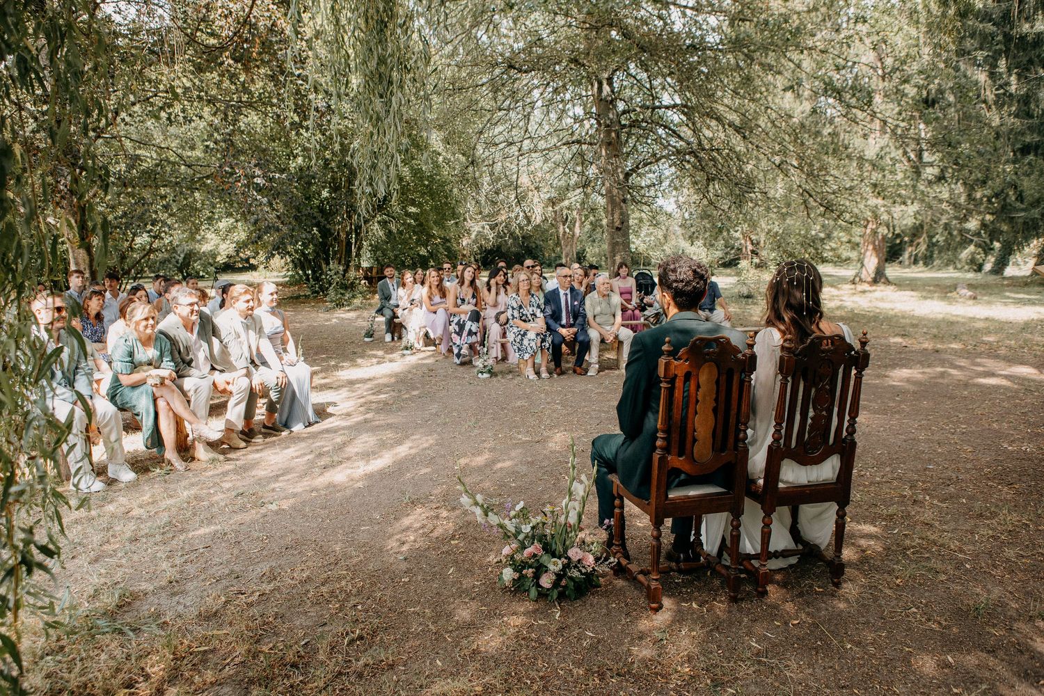 Outdoor wedding ceremony under willow trees with guests seated on benches facing the couple in wooden chairs.