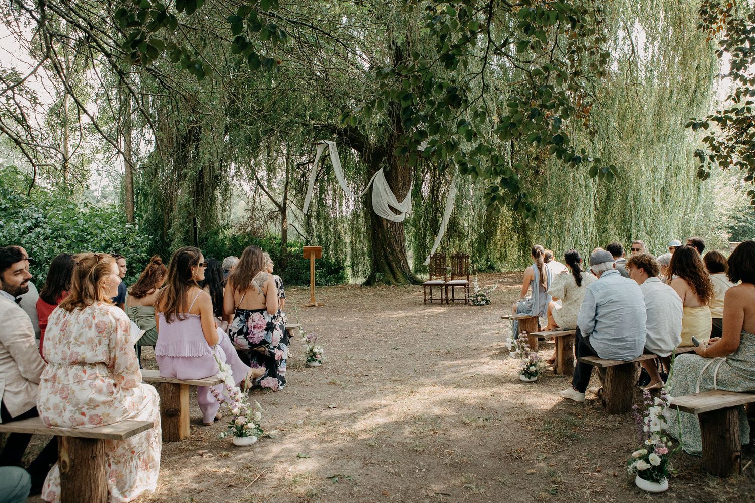 Outdoor wedding ceremony under a large willow tree with guests seated on wooden benches in a natural garden setting.
